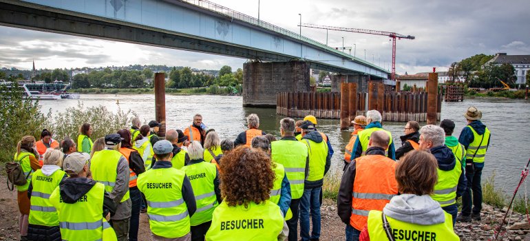 Eine Besuchergruppe steht am Rheinufer. Im Hintergrund sind eine Brücke und Bauarbeiten zu sehen.