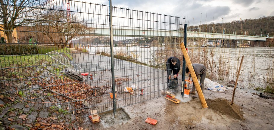 In den Rheinanlagen an der Pfaffendorfer Brücke wird der bisherige Bauzaun derzeit durch einen festen Zaun ersetzt. Seit Beginn des Großbauprojekts war es immer wieder zu Beschädigungen und Zerstörungen am Bauzaun gekommen. Zwei Männer zementieren ein Zaunelement in den Rheinanlagen in Koblenz.