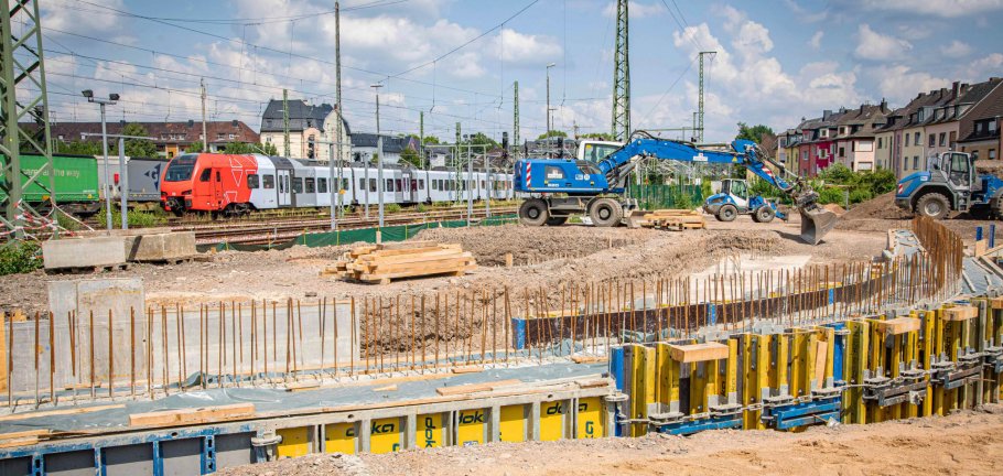 In den kommenden Monaten entsteht zwischen den Koblenzer Stadtteilen Rauental und Goldgrube eine neue Fuß- und Radwegebrücke. Ein Bagger steht an einer Baugrube. Im Hintergrund sind Bahngleise zu sehen.