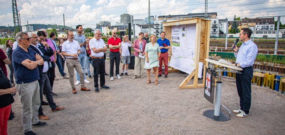 Zum Spatenstich für die neue Geh- und Radwegbrücke zwischen der Goldgrube und Rauental konnte der Koblenzer Oberbürgermeister David Langner (rechts) zahlreiche Gäste aus der Politik sowie von beteiligten Unternehmen begrüßen. Menschen stehen vor einem Rednerpult, an dem ein Mann eine Rede hält.