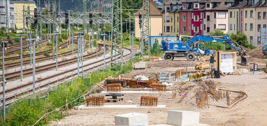 In den kommenden Monaten entsteht zwischen den Koblenzer Stadtteilen Rauental und Goldgrube eine neue Fuß- und Radwegebrücke, die unter anderem über insgesamt zwölf Bahngleise führen wird. Ein Bagger steht an einer Baugrube. Im Hintergrund sind Bahngleise zu sehen.