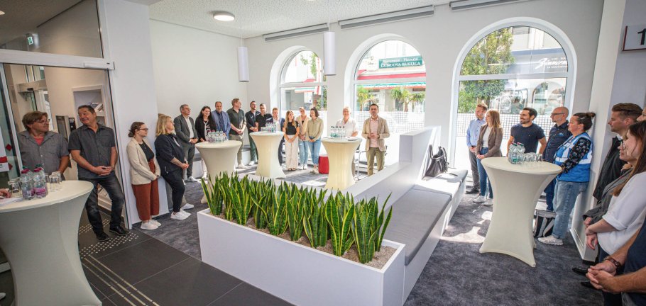Der Koblenzer Oberbürgermeister David Langner und Bürgermeisterin Ulrike Mohrs konnten bei der Eröffnung des umgebauten Bürgeramtes in Koblenz zahlreiche Mitarbeitende sowie beteiligte Ämter und Firmen des Umbaus begrüßen. Menschen stehen in einem lichtdurchfluteten, hellen Raum. Dabei hören sie einer Rede zu.