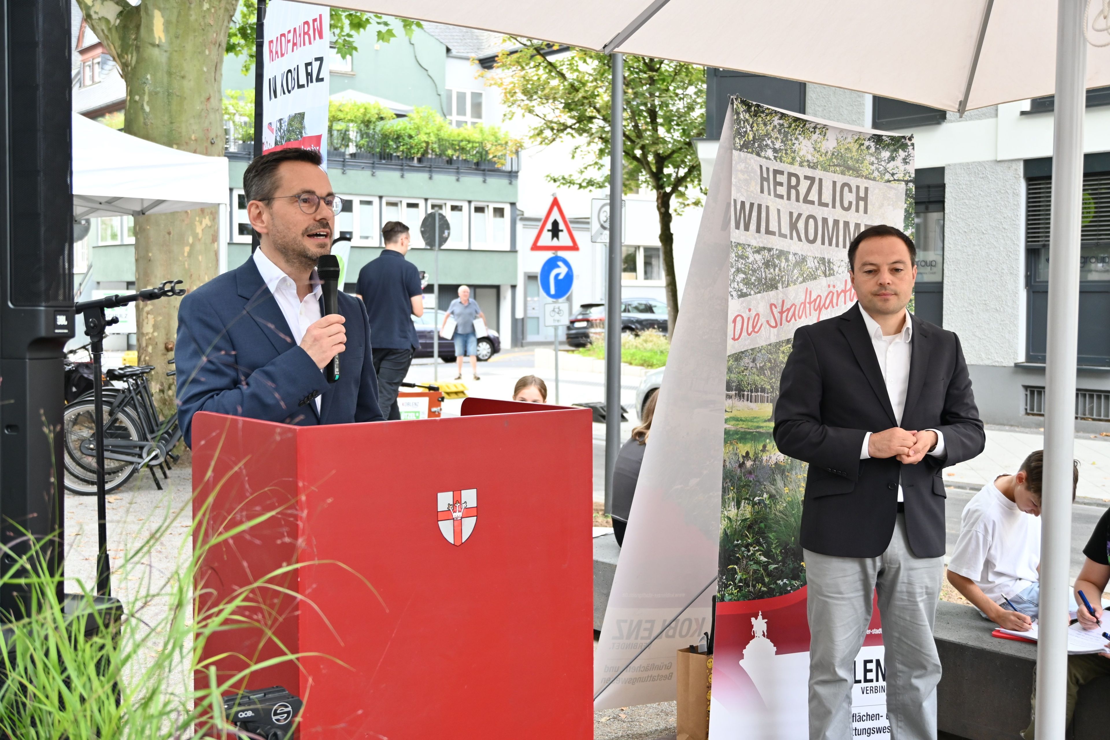 2 Männer stehen unter einem Zeltdach, einer hat ein Mikrophon in der Hand.