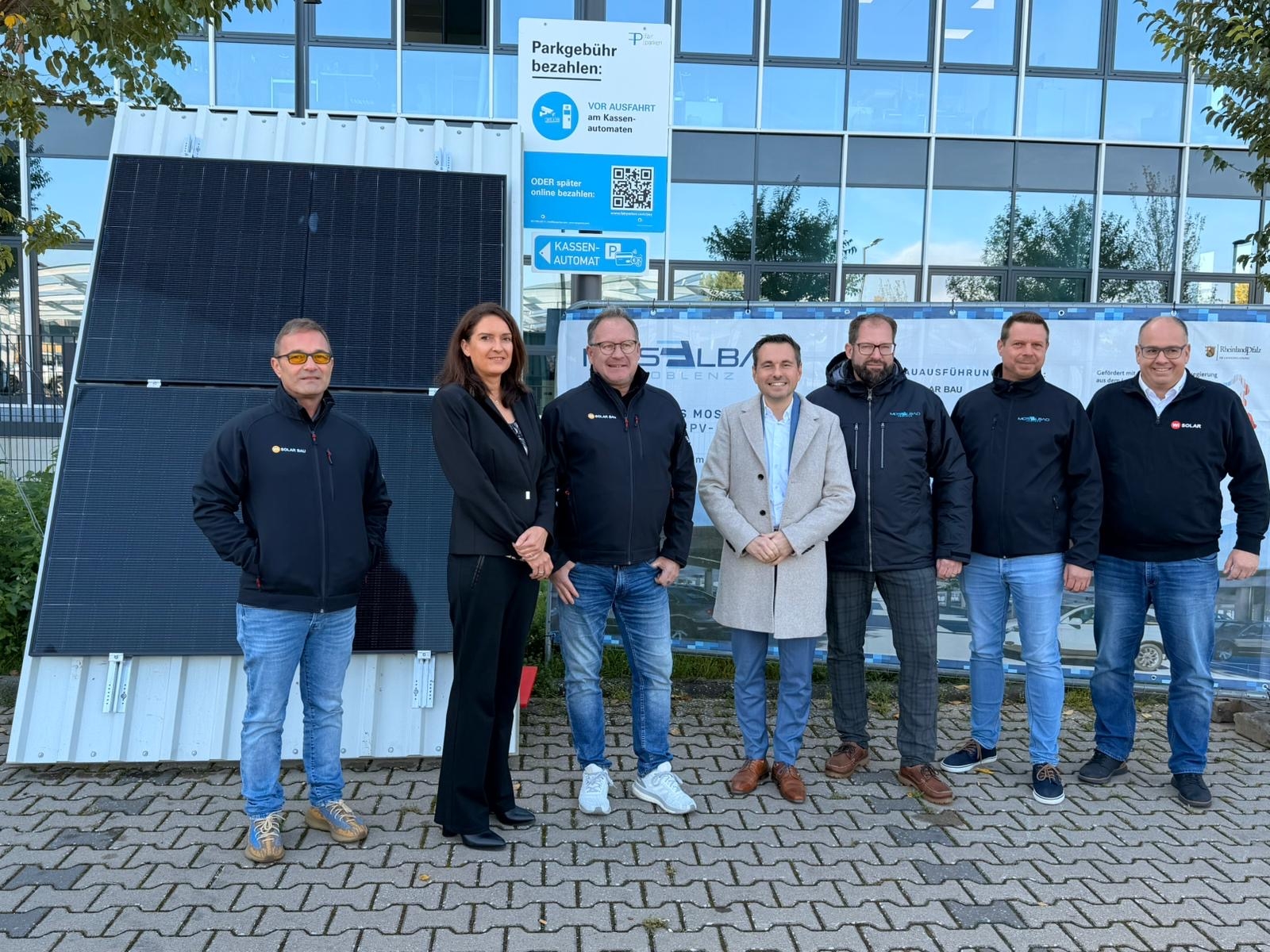 Personen stehen auf einem Parkplatz. Im Hintergrund eine verglaste Gebäudefassade.