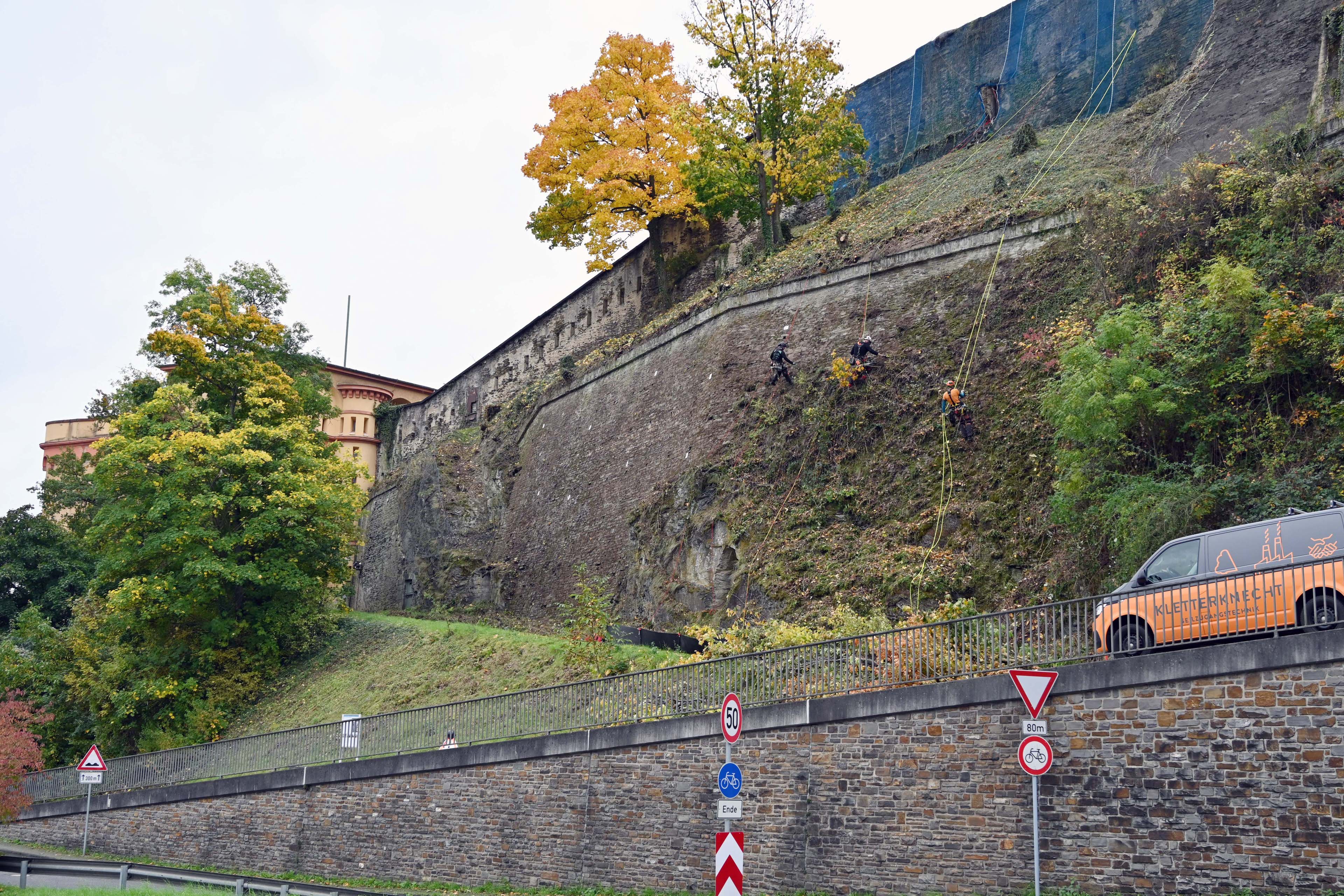 Eine Mauer, an der Männer an Seilen arbeiten. Oberhalb stehen Festungsbauten.