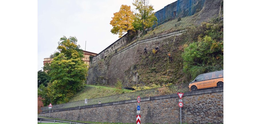 Eine Mauer, an der Männer an Seilen arbeiten. Oberhalb stehen Festungsbauten.