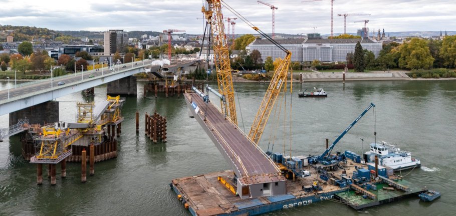 Auch auf der Pfaffendorfer Seite konnten im Oktober die ersten Stahlbauteile für die Pfaffendorfer Brücke per Kran eingehoben werden.  Eine Brückenbaustelle auf dem Rhein aus der Vogelperspektive.