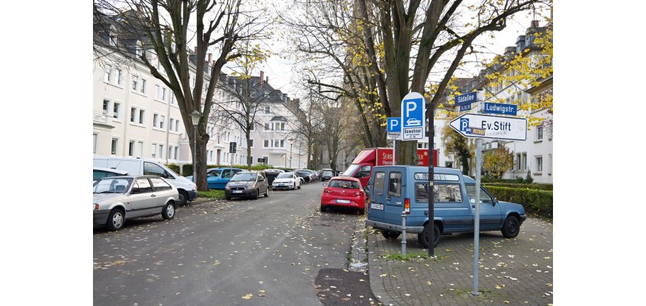 Eine Straße mit parkenden Autos, Bäumen und Hecken vor den Häusern.