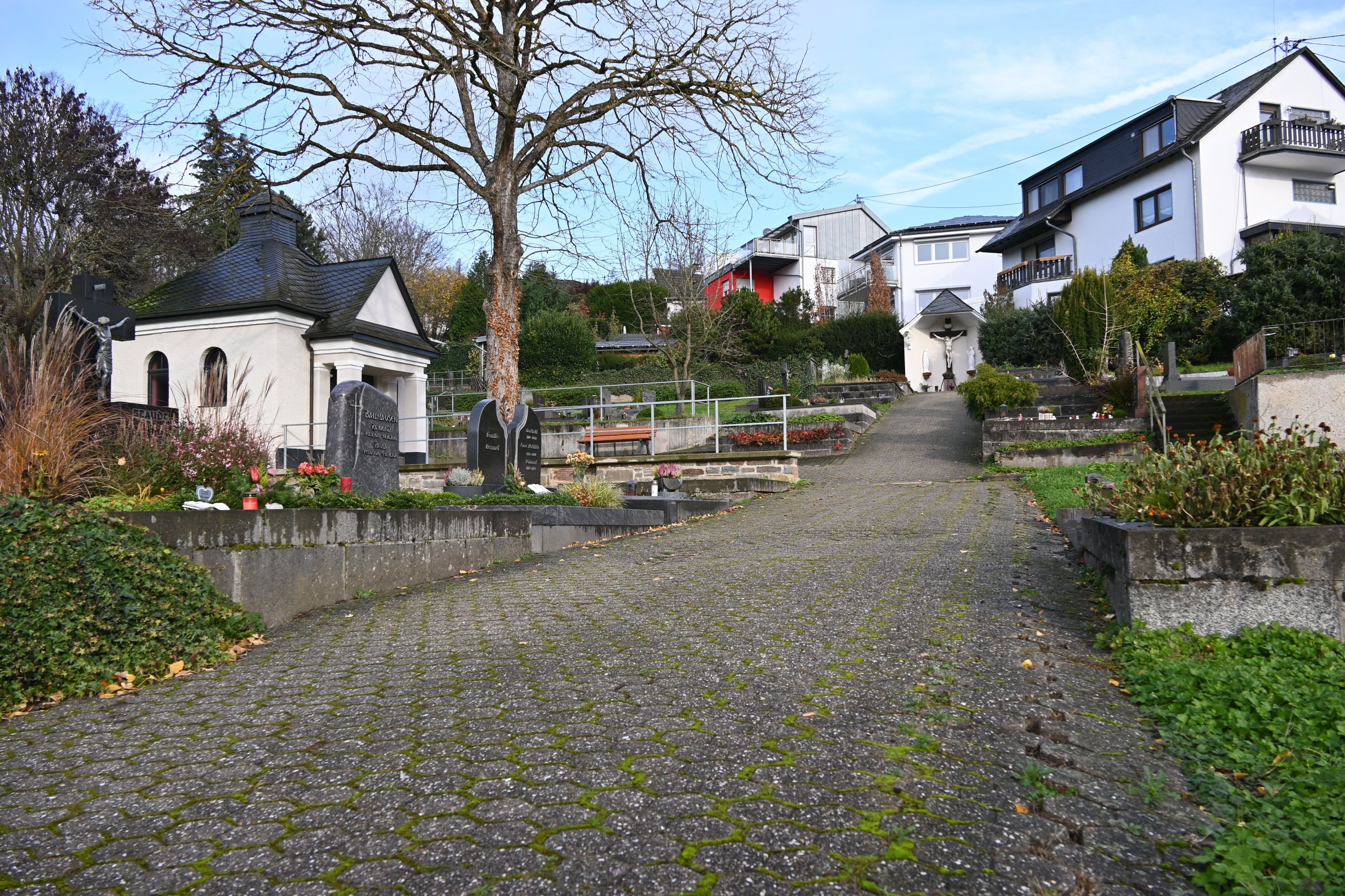 Ein gepflasterter Weg mit sichtbaren Schadstellen mit Gräbern an beiden Seiten. Im Hintergrund steht eine Trauerhalle.