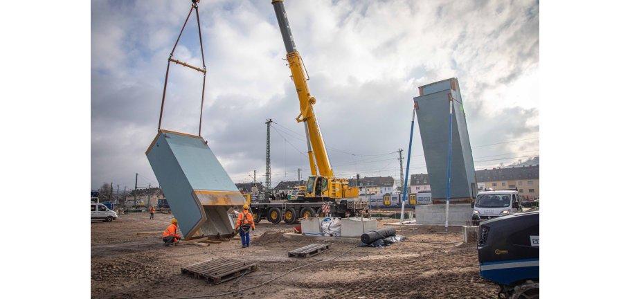 Ein Kran bewegt einen blauen Pfeiler. In der Nähe machen sich Arbeiter in Warnwesten daran zu schaffen.