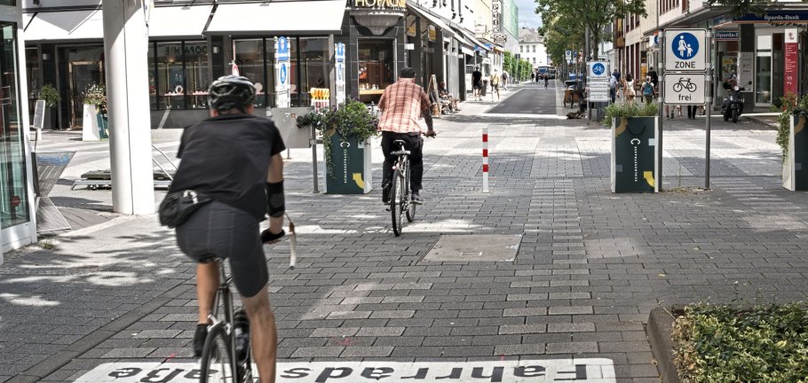 Zwei Radfahrer fahren durch eine Straße. In der Straßenmitte steht ein rot-weißer Roller. Zwei Radfahrer fahren durch eine Straße. In der Straßenmitte steht ein rot-weißer Roller.
